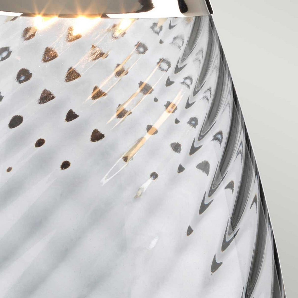 A close-up of a smoke-tinted glass object with wavy patterns and light reflections showcases small dark spots on its surface, complemented by a metallic edge at the top, which emits a warm glow similar to the Lena 2 Light Large Smoke Table Lamp with a Slate Shade. The background is set against a soft, neutral tone.