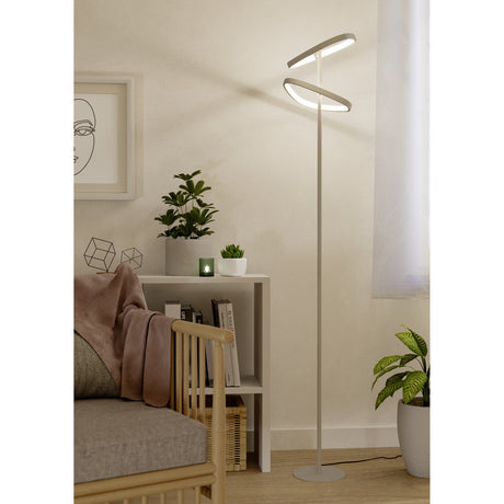Leporano LED Floor Lamp in sand finish with two rounded triangular ring heads on a slim steel pole, shown illuminated in a living room beside plants and a bookshelf.