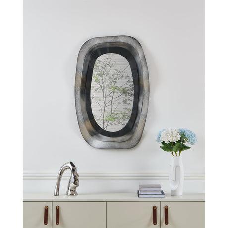 Lircay Wall Decoration, an oval wall mirror with a layered metal frame in brushed silver and black, shown above a cabinet in a light-toned room.