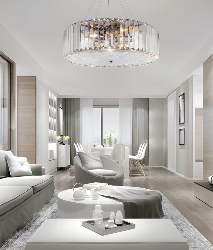 A modern, bright living room with sleek white furniture, featuring a large circular chandelier. A cozy seating area with gray cushions is in the foreground, and a dining table is visible in the background near tall windows.