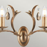 The Lily 2 Light Wall Light - Bronze Patina features a hand-patinated bronze finish with two candle-shaped bulbs. Ornate leaf-like metalwork supports smoked cut glass droplets, offering an elegant and classic design.