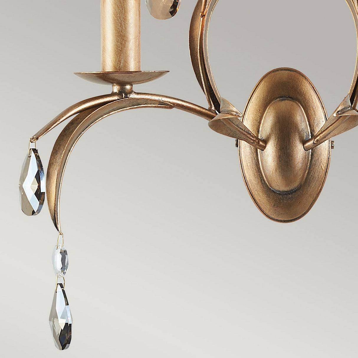 A close-up of an elegant bronze arm from the Lily 2 Light Wall Light - Bronze Patina features oval detailing. Two teardrop-shaped smoked cut glass droplets dangle from the arm, beautifully reflecting light. The neutral grey background perfectly complements the hand-patinated bronze finish characteristic of this design.