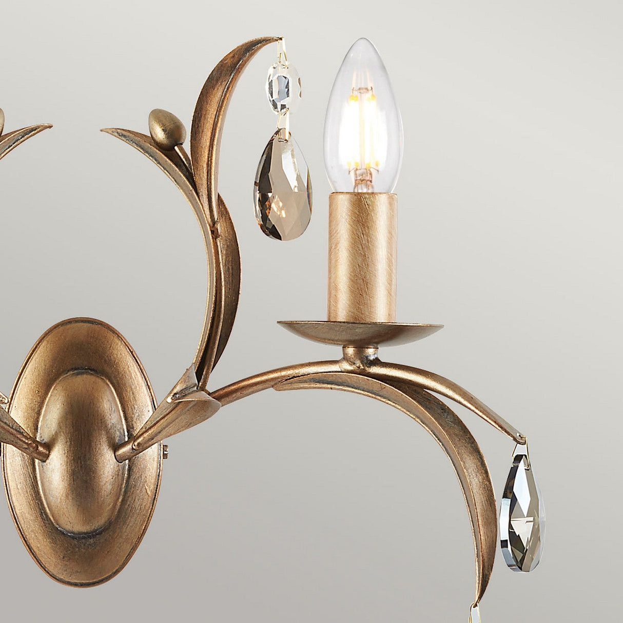 The Lily 2 Light Wall Light - Bronze Patina features a vintage-inspired design with a hand-patinated bronze finish, adorned with decorative crystal pendants. Its unshaded, candle-style bulbs are enhanced by smoked cut glass droplets and graceful, leaf-like curves.