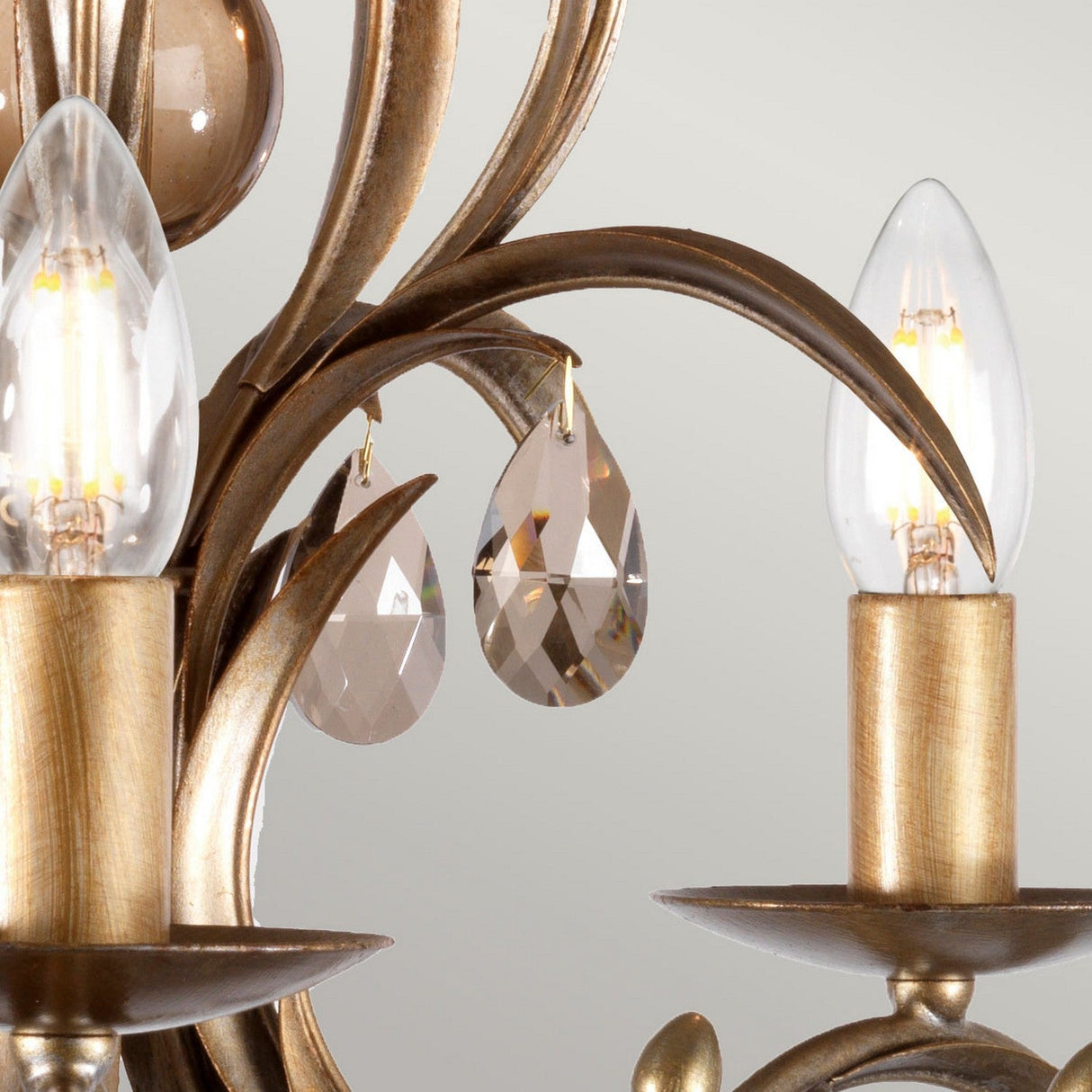 Here's a rewritten version using the provided product data:

A close-up of the Lily 5 Light Chandelier - Bronze Patina reveals its elegant bronze finish, showcasing candle-shaped LED bulbs and smoked cut glass droplets that resemble teardrop-shaped crystals, gracefully hanging from decorative metal curves against a light background.