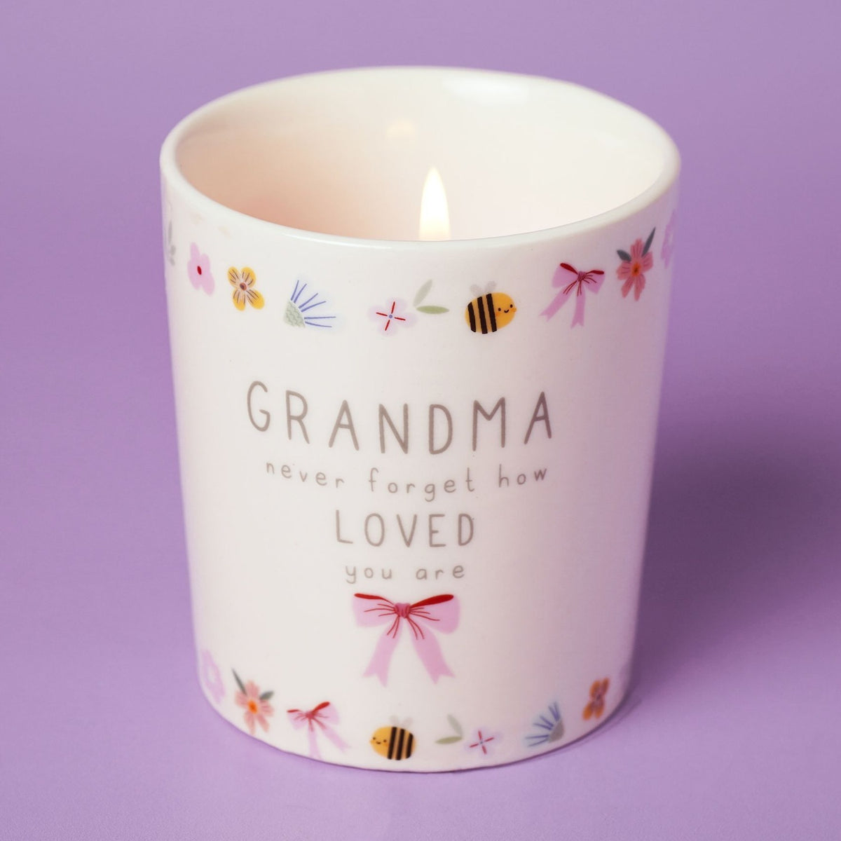 Lit round ceramic scented candle in off-white with "GRANDMA never forget how LOVED you are" text, pink bow, and floral bee border, on a lilac background.