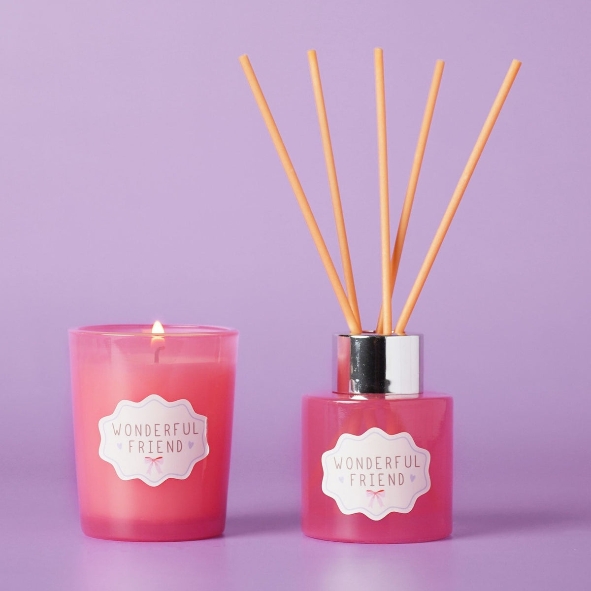 Wonderful Friend Mini Candle and Reed Diffuser Set in pink, with a lit tumbler-style candle and a reed diffuser bottle with silver cap and wooden reeds, both labelled, set against a lilac background.