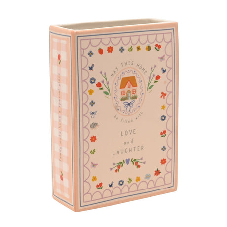 Peach stoneware book-shaped vase printed with "May This Home be Filled With Love and Laughter" around a cottage, with a floral scalloped border and gingham spine, on a white background.