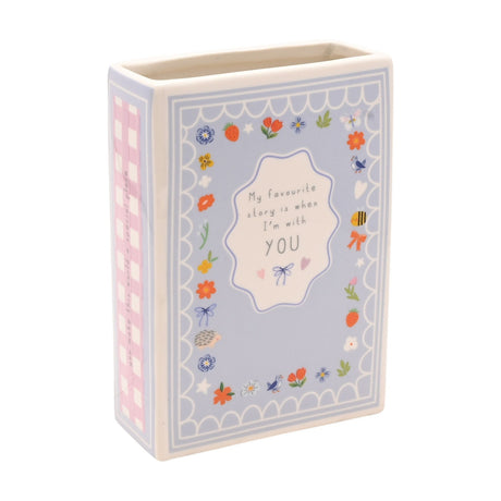 Blue stoneware book-shaped vase with floral, bee, and bird border around "My favourite story is when I'm with YOU" text, pink gingham spine, on a white background.