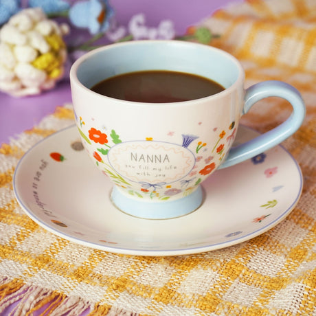 Off white stoneware cup with light blue interior and handle, filled with coffee, printed with "NANNA, you fill my life with joy" amid floral designs, on a matching saucer.