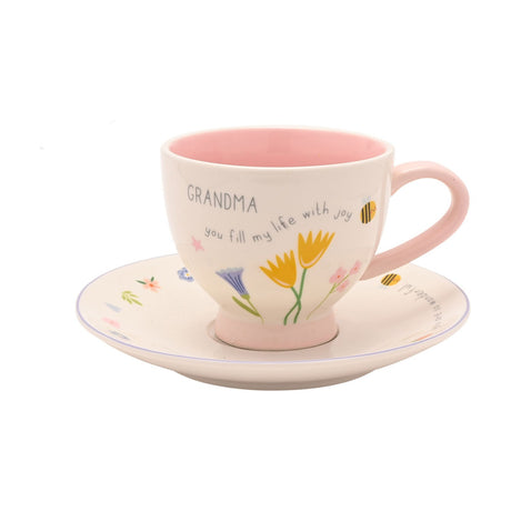 Stoneware cup and saucer with "GRANDMA you fill my life with joy" text, pink interior and handle, floral tulip design, on a plain white background.