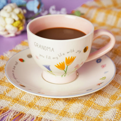 Grandma Floral Stoneware Cup & Saucer in off white with a pink interior and handle, printed with "Grandma, you fill my life with joy" and tulip and bellflower florals, filled with hot chocolate.