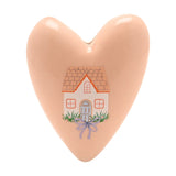 Pink stoneware heart-shaped ornament with a painted cottage design featuring a tiled roof, windows, arched door, and lilac bow, set against a plain white background.