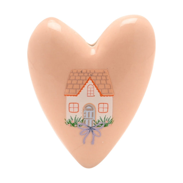 Pink stoneware heart-shaped ornament with a painted cottage design featuring a tiled roof, windows, arched door, and lilac bow, set against a plain white background.
