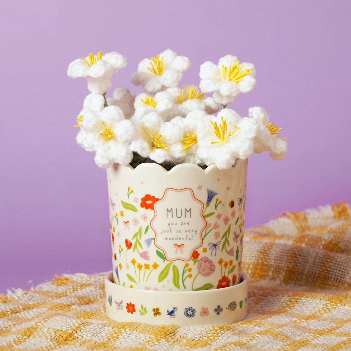 Off-white stoneware planter with scalloped rim and floral, bee and butterfly print, framed "MUM" message, matching saucer, holding white crocheted flowers on a lilac background.