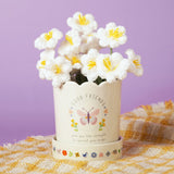 Off-white stoneware planter with scalloped rim, Good Friends text, and butterfly decal, shown with matching saucer, crochet flowers, and plaid cloth on a lilac background.