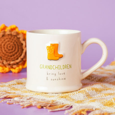 Cream stoneware mug with embossed yellow and orange welly boots above "GRANDCHILDREN bring love & sunshine" text, shown on a yellow woven throw against a lilac background.
