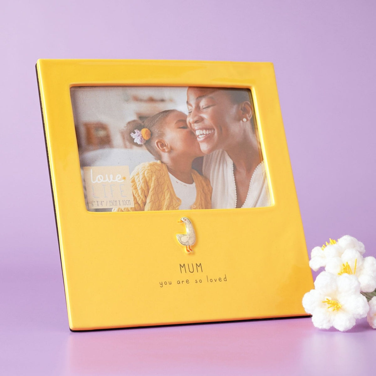 Yellow glazed stoneware photo frame holding a 6 x 4in landscape photo of a mother and child, with a raised goose detail and "MUM you are so loved" text below, shown against a lilac background.
