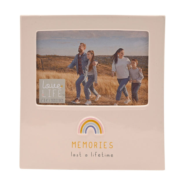 Cream glazed stoneware photo frame with rectangular 6 x 4in window, pastel rainbow detail and "Memories last a lifetime" text below, shown with a family walking photo on a plain white background.