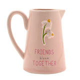 Pink stoneware jug with curved handle, featuring embossed white flowers with yellow centres and "Friends bloom together" text, shown on a plain white background.