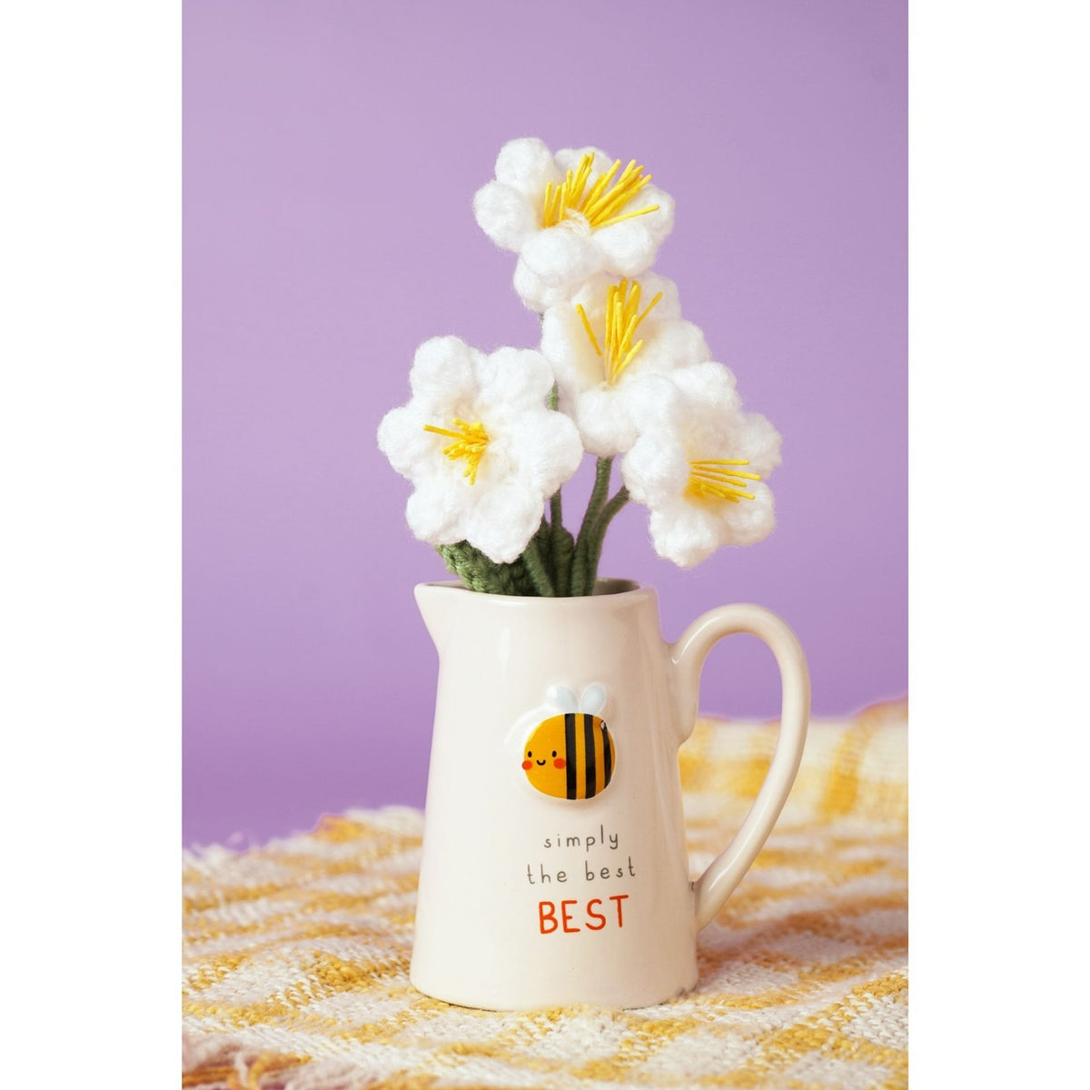 Cream stoneware jug with embossed smiling bee design and "simply the BEST" text, shown holding white crocheted flowers on a yellow plaid cloth against a purple background.
