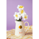 Cream stoneware jug with embossed smiling bee design and "simply the BEST" text, shown holding white crocheted flowers on a yellow plaid cloth against a purple background.