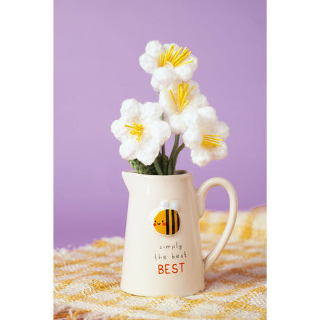 Cream stoneware jug with embossed smiling bee design and "simply the BEST" text, shown holding white crocheted flowers on a yellow plaid cloth against a purple background.