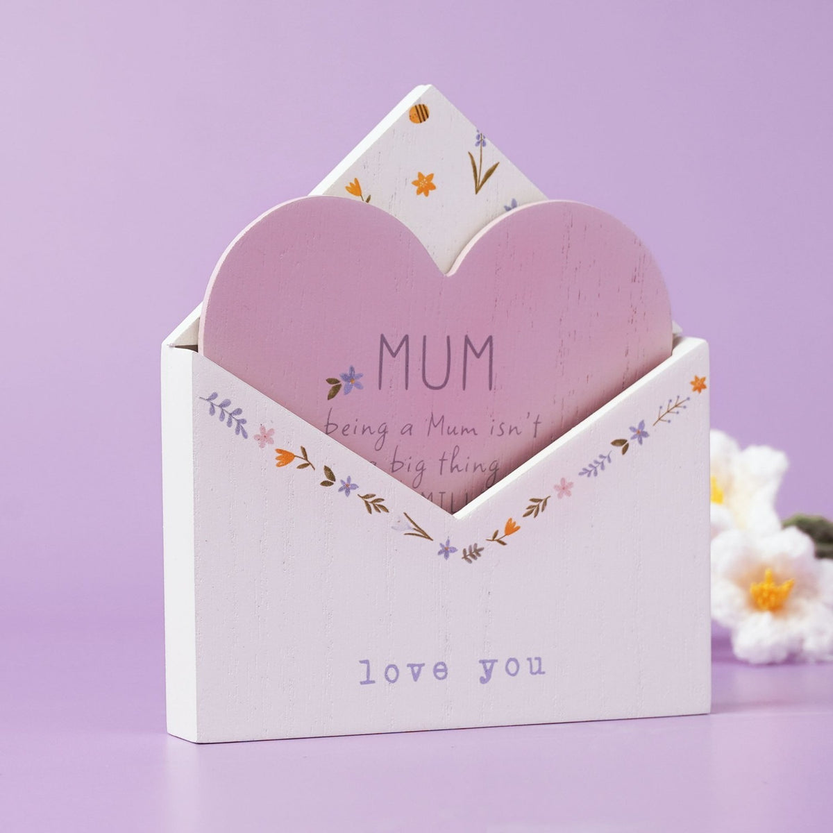 White wooden envelope plaque with pink heart insert reading "MUM", floral border, and "love you" text, set against a lilac background with faux flowers.