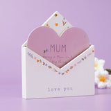White wooden envelope plaque with pink heart insert reading "MUM", floral border, and "love you" text, set against a lilac background with faux flowers.