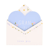 White wooden envelope-shaped wall plaque with a pale blue heart insert reading "GRAN," floral details in pink, yellow and lilac, on a plain white background.