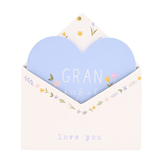 White wooden envelope-shaped wall plaque with a pale blue heart insert reading "GRAN," floral details in pink, yellow and lilac, on a plain white background.