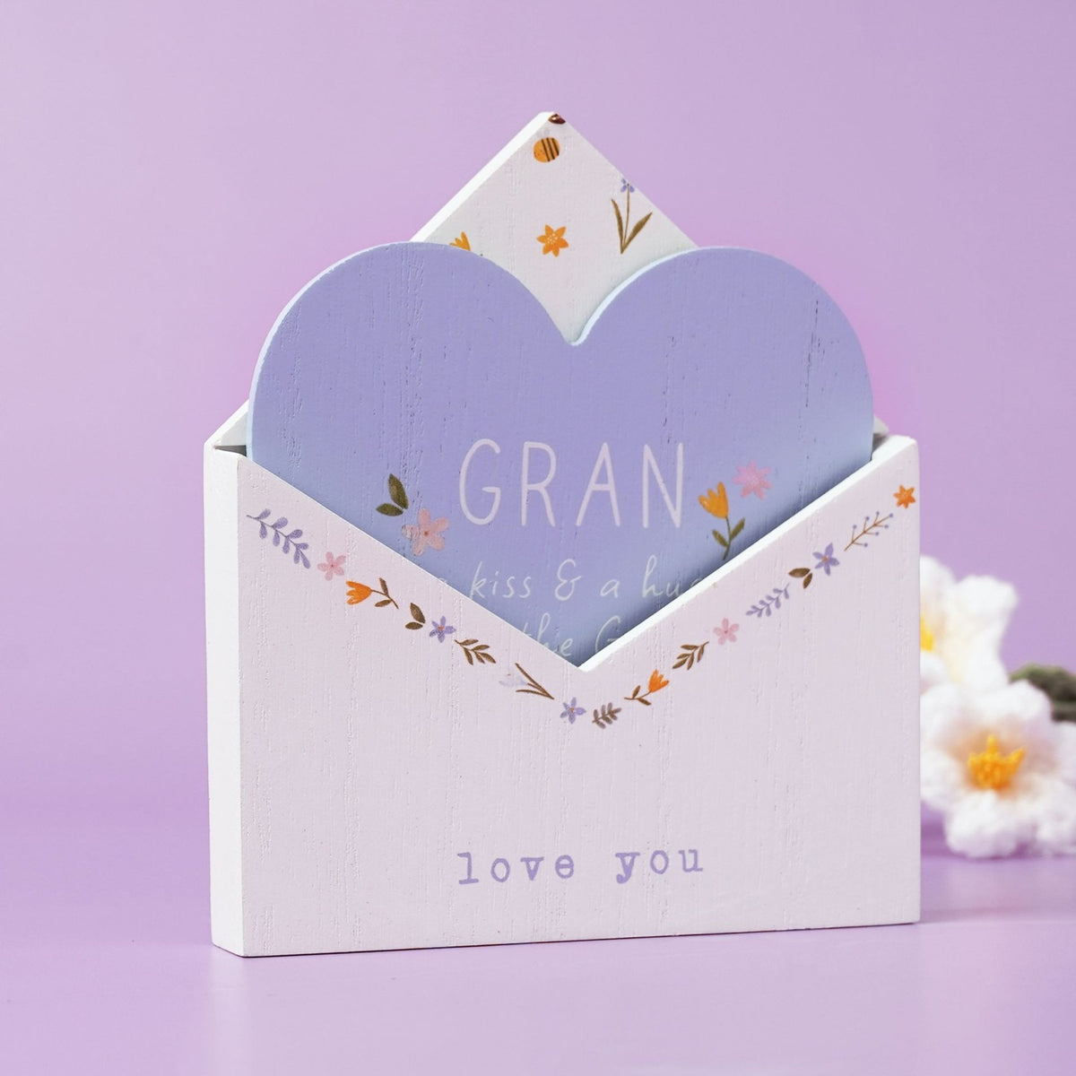 White wooden envelope plaque with a pale blue heart insert reading "GRAN", floral details in pink, yellow and lilac, and "love you" text, on a lilac background.