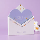 White wooden envelope plaque with a pale blue heart insert reading "GRAN", floral details in pink, yellow and lilac, and "love you" text, on a lilac background.