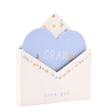 White wooden envelope plaque with a pale blue heart insert reading "GRAN," floral details in pink, yellow and lilac, and "love you" text below, shown on a plain white background.