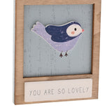 Wooden wall plaque featuring a raised lilac and navy bird with white face and pink beak on a weathered blue panel, framed in natural wood with "You Are So Lovely" in grey capitals below.
