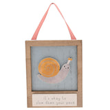 Wooden wall plaque featuring a raised orange and grey snail on a weathered pale blue panel with natural wood frame, cream base reading it's okay to slow down your pace, and pink ribbon hanger.