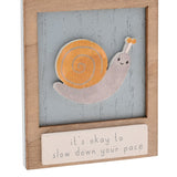 Wooden wall plaque featuring a raised smiling snail with orange spiral shell on a weathered blue panel, framed in natural wood with a cream message strip reading "it's okay to slow down your pace."