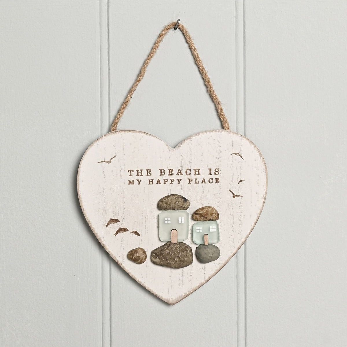 White heart-shaped wall plaque with whitewashed grain finish reading THE BEACH IS MY HAPPY PLACE, featuring two frosted green glass cottages with bird motifs on pebble bases, hung on jute cord.