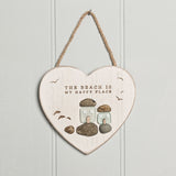White heart-shaped wall plaque with whitewashed grain finish reading THE BEACH IS MY HAPPY PLACE, featuring two frosted green glass cottages with bird motifs on pebble bases, hung on jute cord.