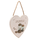 White heart-shaped wall plaque with a whitewashed finish, jute hanging cord, "THE BEACH IS MY HAPPY PLACE" text, engraved gulls, and two frosted green glass cottages on pebble shapes.