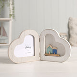 White wooden folding double heart photo frame with a heart-shaped photo window on one side and a shallow alcove displaying two small sea-green and blue houses on the other.