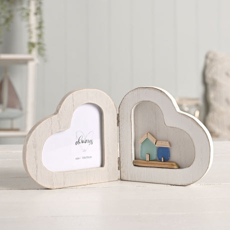 White wooden folding double heart photo frame with a heart-shaped photo window on one side and a shallow alcove displaying two small sea-green and blue houses on the other.
