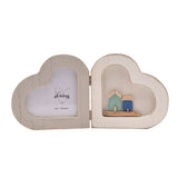 Folding wooden double heart photo frame in white, one side with a heart-shaped photo window, the other framing a small seaside scene with green and blue houses on a wooden base.