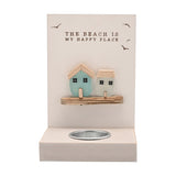 Cream tea light candle holder with sea glass houses in aqua and frosted white, "THE BEACH IS MY HAPPY PLACE" text and bird motifs on a wood plaque above a metal tea light cup, plain background.
