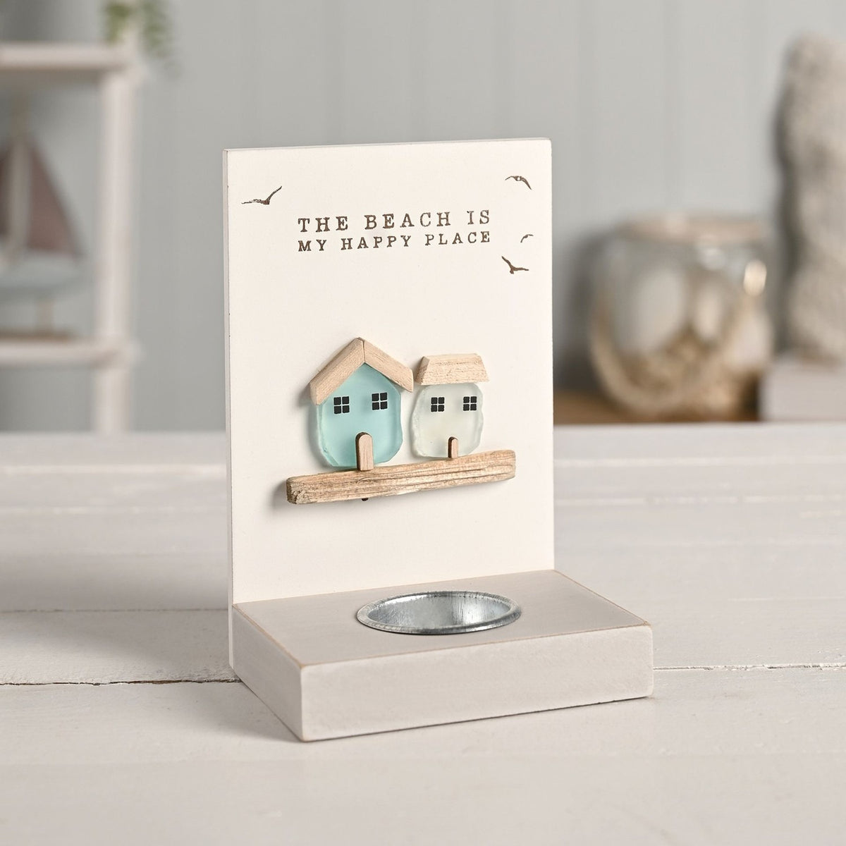 Cream tea light candle holder with two sea glass houses, aqua and frosted white, on a wooden ledge beneath "The Beach Is My Happy Place" text and bird motifs, set on a light tabletop.