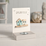 Cream tea light candle holder with two sea glass houses, aqua and frosted white, on a wooden ledge beneath "The Beach Is My Happy Place" text and bird motifs, set on a light tabletop.