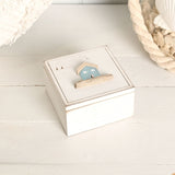 Square white wooden trinket box with hinged lid, featuring a pale blue sea glass house motif, driftwood-style base, and engraved birds and trees on the lid.
