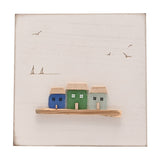 White square wall plaque with three sea glass cottages (blue, green, aqua) on a driftwood ledge, with engraved seagulls and sailing boats on a plain white background.