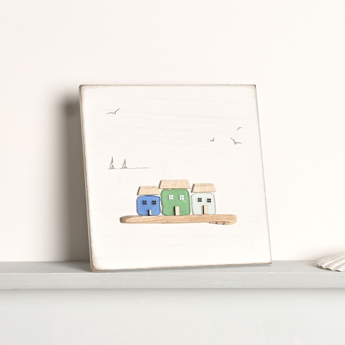 By the Sea Square Plaque with 3 Glass Houses in blue, green and aqua, white wood background with gull and boat line drawings, leaning on a shelf.