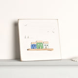 By the Sea Square Plaque with 3 Glass Houses in blue, green and aqua, white wood background with gull and boat line drawings, leaning on a shelf.