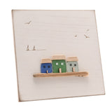 White square wall plaque with three sea glass houses in blue, green and aqua on a driftwood ledge, etched gulls and boats on a white background.